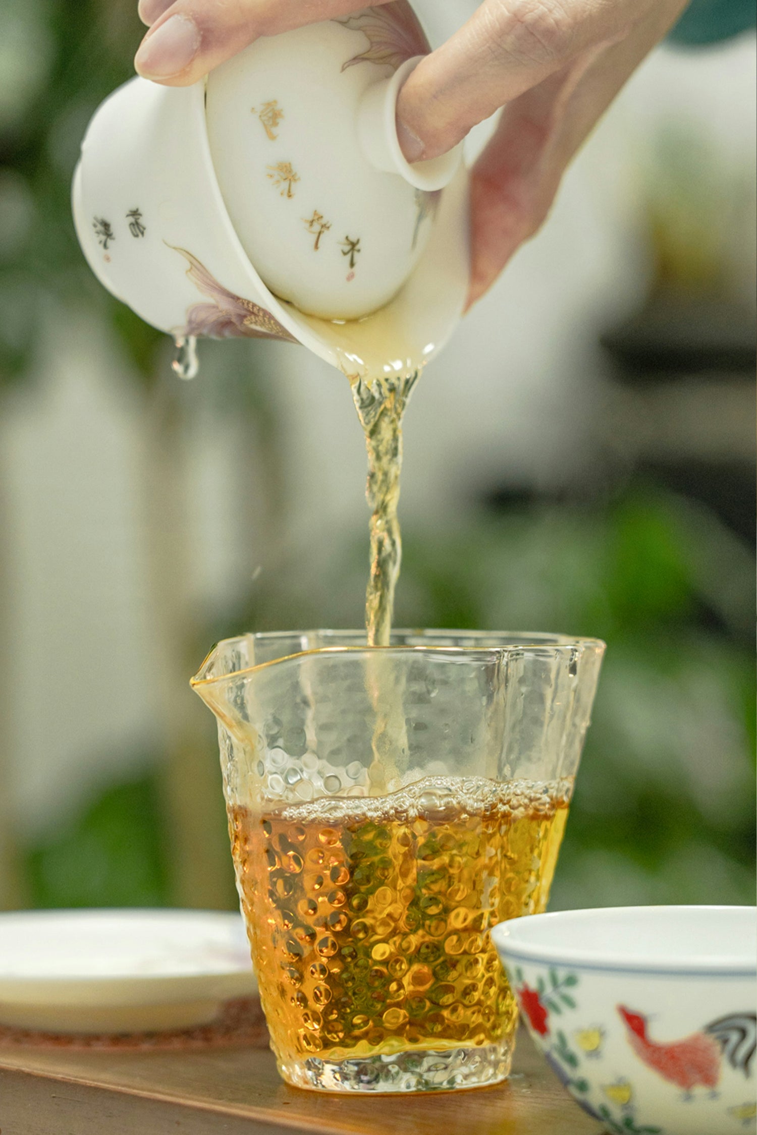 Tea being poured from a teapot into a glass cup with a blurred natural background