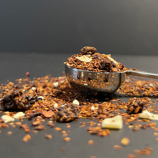 A luxurious close-up of Cocoa Forest Bloom loose leaf tea, showing dark black currants and white chocolate curls nestled among rich red Rooibos and Honeybush leaves.